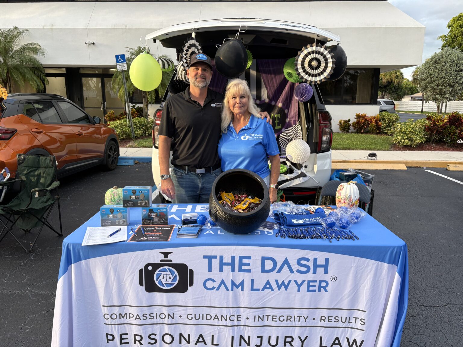 Shannon Sagan in a black polo and cap and Joy in a blue The Dash Cam Lawyer polo stand behind a branded blue-and-white table with a cauldron of candy and dash cam boxes, in front of an SUV trunk decorated in black, white, purple and green.