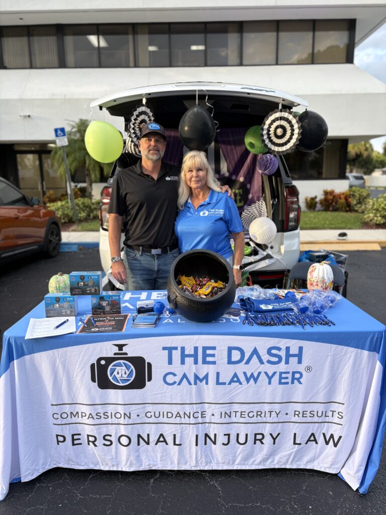 Shannon Sagan in a black polo and cap and Joy in a blue The Dash Cam Lawyer polo stand behind a branded blue-and-white table with a cauldron of candy and dash cam boxes, in front of an SUV trunk decorated in black, white, purple and green.