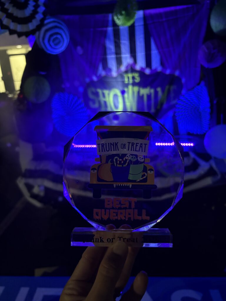 A hand holds a clear acrylic Trunk or Treat Best Overall award in front of the decorated trunk glowing under black light.